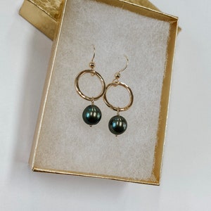 May include: A pair of gold hoop earrings with a black pearl dangling from each hoop. The earrings are displayed in a white gift box with a gold trim.
