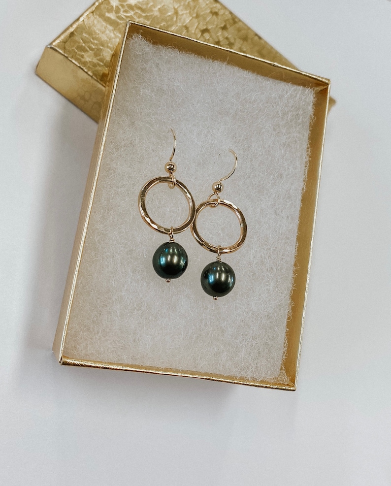 May include: A pair of gold hoop earrings with a black pearl dangling from each hoop. The earrings are displayed in a white gift box with a gold trim.