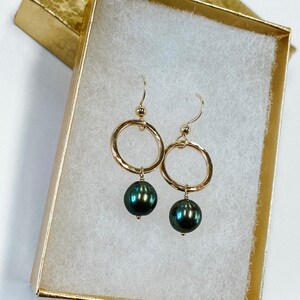 May include: A pair of gold hoop earrings with a black pearl dangling from each hoop. The earrings are in a white box with a gold lid.