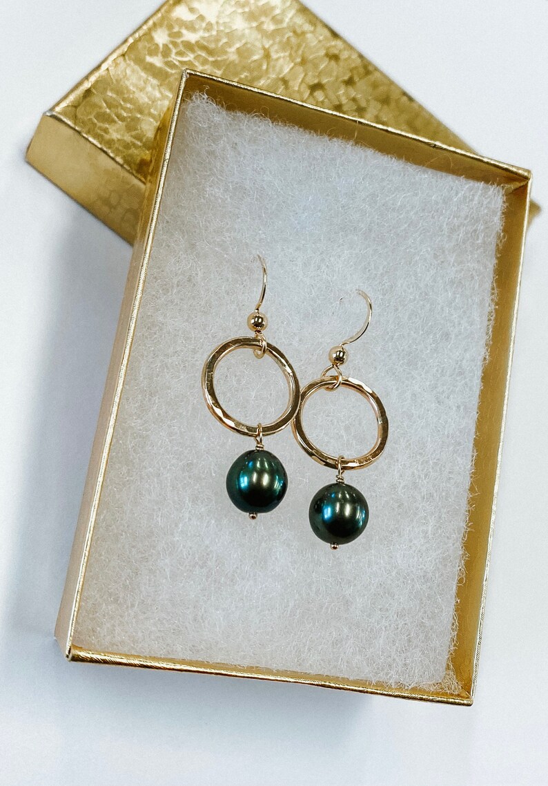 May include: A pair of gold hoop earrings with a black pearl dangling from each hoop. The earrings are in a white box with a gold lid.