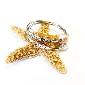 Puede incluir: Dos pulseras de plata con los nombres "Ki-Lyn" y "Yukiko" grabados en ellas. Las pulseras est&aacute;n apiladas sobre una estrella de mar y una perla rosa est&aacute; unida a la pulsera superior.