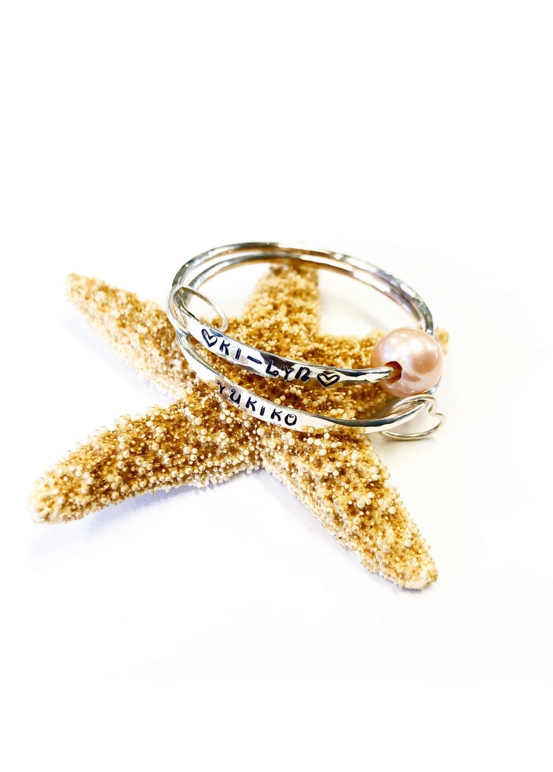 Puede incluir: Dos pulseras de plata con los nombres "Ki-Lyn" y "Yukiko" grabados en ellas. Las pulseras est&aacute;n apiladas sobre una estrella de mar y una perla rosa est&aacute; unida a la pulsera superior.
