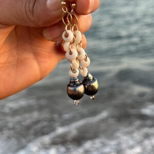 May include: Gold-tone earrings with a dangling design. Each earring features a series of small, white, circular beads, leading to a dark gray, pear-shaped bead with a small, clear gem at the bottom. The earrings are held up by gold-colored hooks.