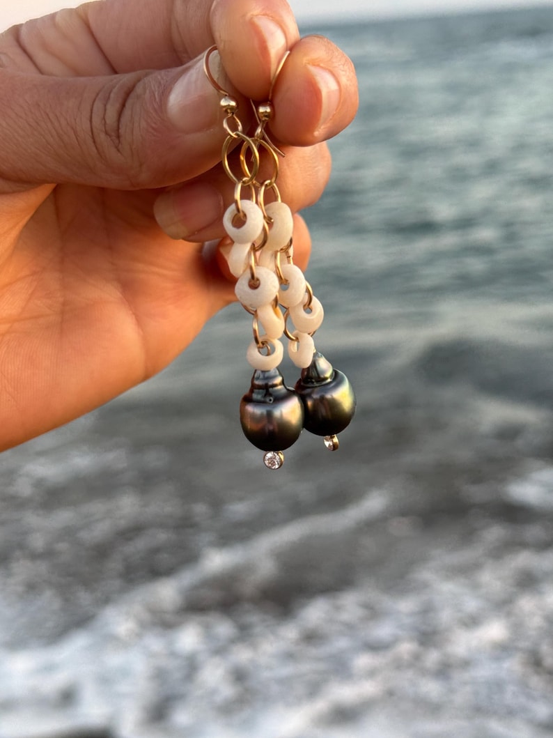 May include: Gold-tone earrings with a dangling design. Each earring features a series of small, white, circular beads, leading to a dark gray, pear-shaped bead with a small, clear gem at the bottom. The earrings are held up by gold-colored hooks.