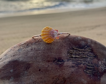 May include: A gold-toned metal bangle bracelet featuring a single, small, fan-shaped yellow and orange seashell attached to the centre. The shell is secured to the thin, polished metal band with delicate gold-coloured wire wrapping.