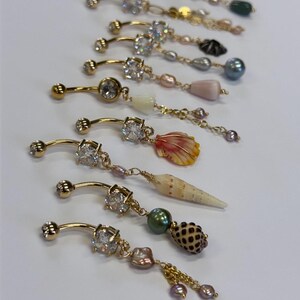 May include: A collection of gold-toned navel rings featuring clear cubic zirconia stones and dangling charms. The charms include various natural seashells, freshwater pearls, and small metallic beads attached to delicate gold-colored chains.