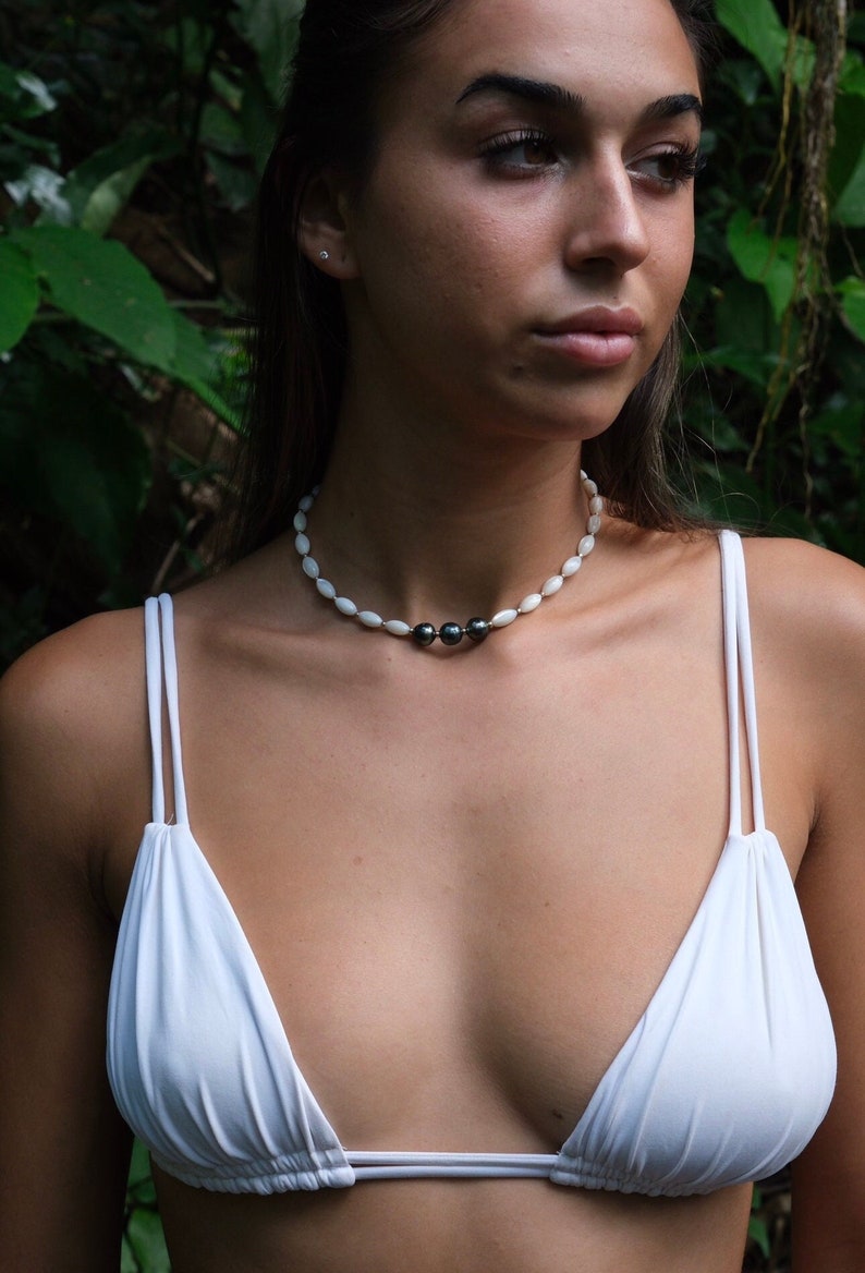 Op de afbeelding: Een vrouw draagt een witte bikini top met een witte en zwarte parelketting. De ketting heeft drie zwarte parels in het midden.