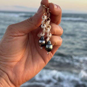 May include: Gold-toned dangle earrings featuring dark gray pearls and white shell accents. The earrings have a delicate design with small, dangling shells and a single, sparkling accent at the base of each pearl. The background shows the ocean.