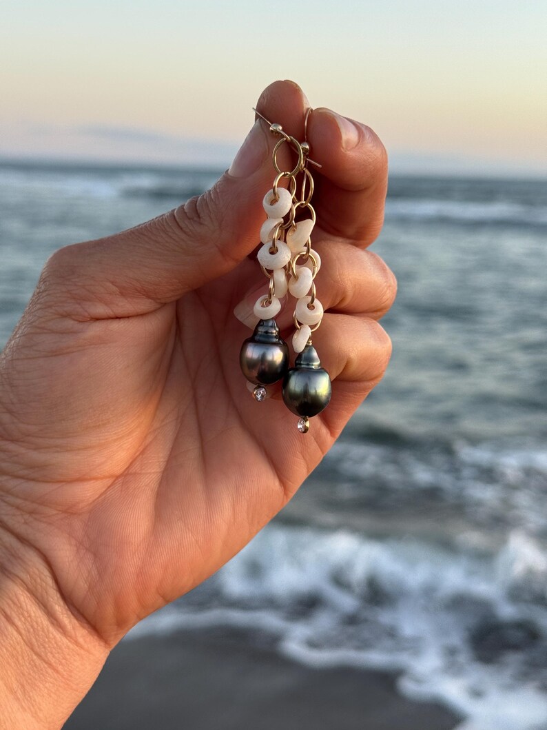May include: Gold-toned dangle earrings featuring dark gray pearls and white shell accents. The earrings have a delicate design with small, dangling shells and a single, sparkling accent at the base of each pearl. The background shows the ocean.