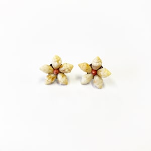 May include: A pair of flower-shaped earrings. Each earring is composed of five small, off-white seashells arranged around a central, reddish-orange bead. The earrings are set on a dark brown base, creating a natural, beachy aesthetic.