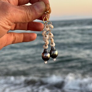 May include: Gold-toned earrings featuring a cascade of small, white shell-like beads and two dark gray, pear-shaped pearls. Each pearl is accented with a small, sparkling clear stone. The earrings are held against a blurred ocean backdrop.