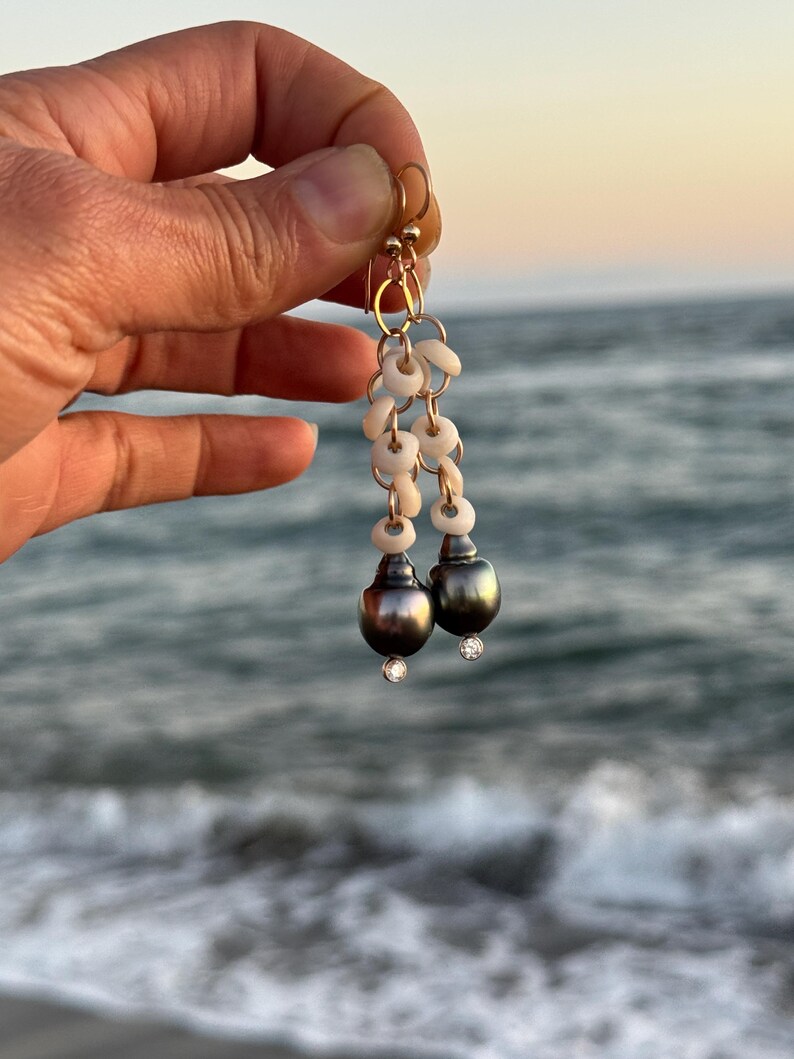 May include: Gold-toned earrings featuring a cascade of small, white shell-like beads and two dark gray, pear-shaped pearls. Each pearl is accented with a small, sparkling clear stone. The earrings are held against a blurred ocean backdrop.