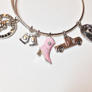 May include: A silver charm bracelet with multiple charms, including a cowgirl, denim shorts, a pink boot, a pickup truck, and a cowboy hat.