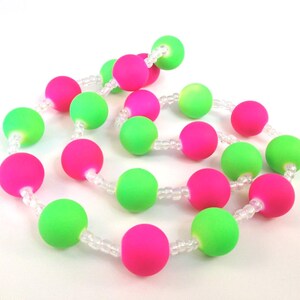 May include: A necklace featuring alternating matte neon pink and neon green spherical beads strung together with small, clear, translucent spacer beads. The beads are arranged in a continuous, coiled pattern against a plain white background.