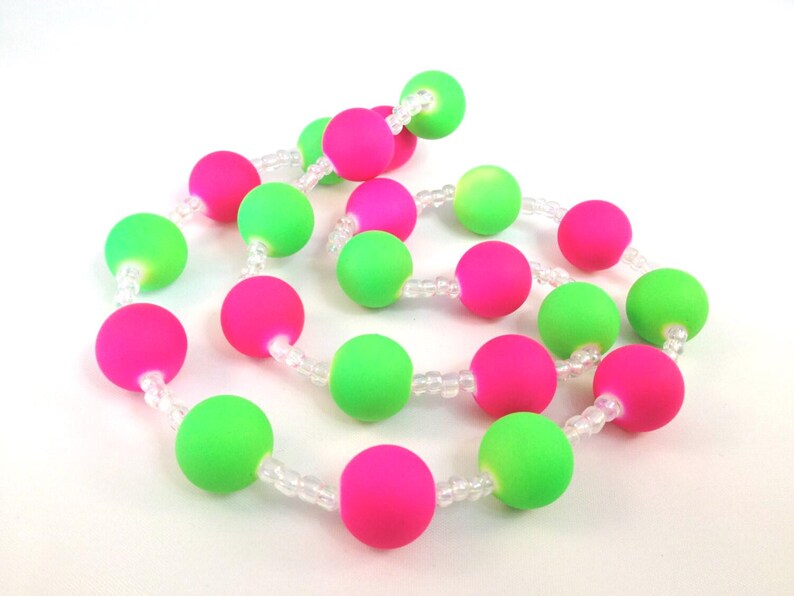 May include: A necklace featuring alternating matte neon pink and neon green spherical beads strung together with small, clear, translucent spacer beads. The beads are arranged in a continuous, coiled pattern against a plain white background.