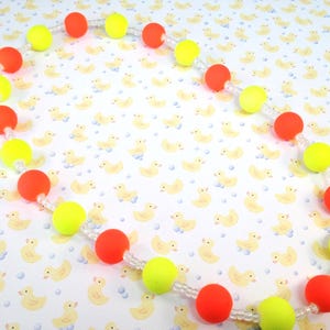 May include: A necklace featuring alternating round neon orange and neon yellow beads strung on a clear cord. The beads are arranged in a repeating pattern, separated by small clear spacer beads, creating a vibrant and colorful circular design.