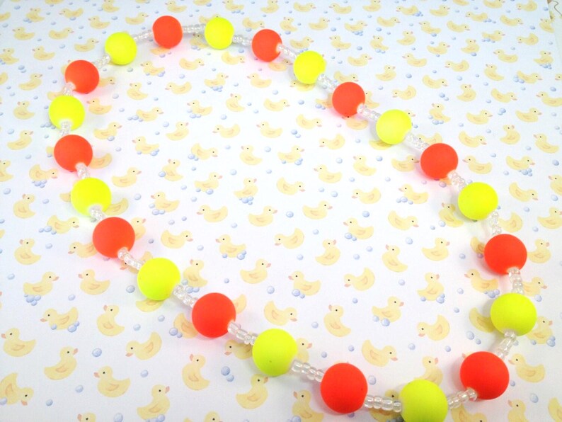 May include: A necklace featuring alternating round neon orange and neon yellow beads strung on a clear cord. The beads are arranged in a repeating pattern, separated by small clear spacer beads, creating a vibrant and colorful circular design.