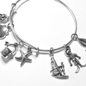 May include: A silver-toned metal charm bracelet featuring seven distinct nautical-themed charms: a pair of flip-flops, a seashell, a sand bucket with the word SAND embossed on it, a starfish, a windsurfer, a scuba diver, and a sailboat.
