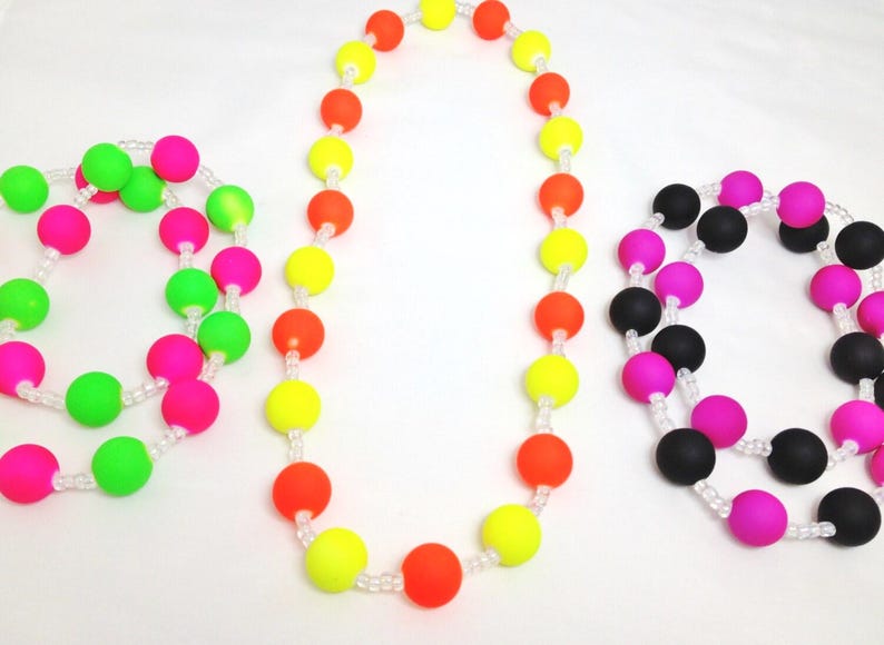 May include: Three beaded necklaces featuring round, matte-finish silicone beads strung on clear cord. One necklace has neon green and pink beads, one has neon yellow and orange beads, and one has alternating black and bright pink beads.