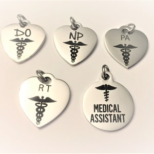May include: Five silver medical ID tags with a caduceus symbol and different medical abbreviations. The tags are in the shape of a heart except for one which is round. The round tag says "Medical Assistant".