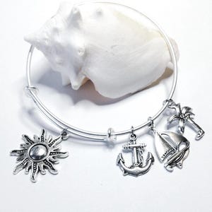 May include: A silver-toned expandable bangle bracelet featuring four dangling charms: a sun, an anchor, a sailboat, and a palm tree. The bracelet is positioned around a large, white, textured seashell, showcasing a nautical and coastal theme.