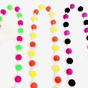 May include: Three beaded necklaces arranged vertically. The first features alternating neon pink and lime green spherical beads. The second displays alternating bright yellow and orange beads. The third consists of alternating black and magenta beads.