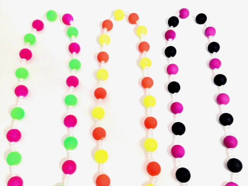 May include: Three beaded necklaces arranged vertically. The first features alternating neon pink and lime green spherical beads. The second displays alternating bright yellow and orange beads. The third consists of alternating black and magenta beads.