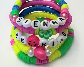 May include: Multicolored beaded bracelets with the words "Sienna" and "Be Kind" spelled out in black letters. The bracelets also feature a smiley face bead and a pink heart bead.