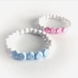May include: Two elastic beaded bracelets featuring white round beads. One bracelet displays the name EMMA in pink letter beads, while the other bracelet displays the name ETHAN in light blue letter beads. The beads are arranged in a circular pattern.