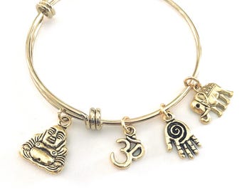 May include: A gold-toned expandable wire bangle bracelet featuring four hanging charms: a laughing Buddha figure, an Om symbol, a hamsa hand with a spiral centre, and an elephant, all finished in a matching metallic gold tone.