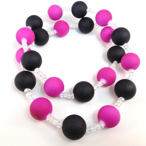 May include: A necklace featuring alternating matte black and vibrant fuchsia spherical beads, strung together with small, clear, translucent spacer beads. The circular design displays a repeating pattern of these colored spheres throughout the strand.