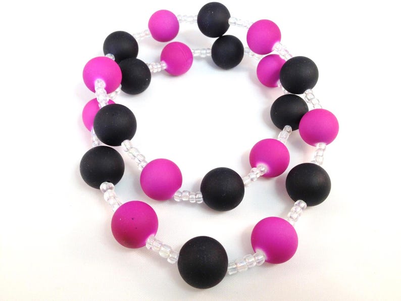 May include: A necklace featuring alternating matte black and vibrant fuchsia spherical beads, strung together with small, clear, translucent spacer beads. The circular design displays a repeating pattern of these colored spheres throughout the strand.