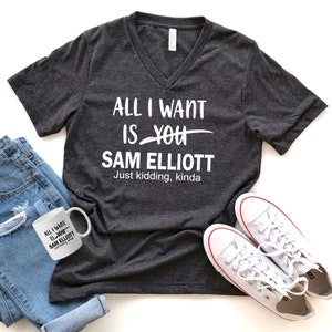 May include: A dark gray V-neck t-shirt with the text "ALL I WANT IS YOU SAM ELLIOTT Just kidding, kinda" printed in white.