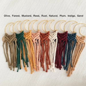 May include: Ten macrame wall hangings in various colors, including olive, forest green, mustard yellow, rose, rust, natural, plum, indigo, and sand. Each hanging features a crescent moon shape and a tassel fringe.