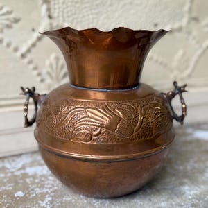 May include: A copper-colored vase with a wide, ruffled rim and a bulbous body. The body features an embossed floral design and two ornate handles. The vase has a vintage look and is likely made of solid copper.