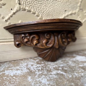 May include: A semi-circular wall shelf crafted from dark brown wood, featuring a flat top surface and a decorative support bracket adorned with intricate carved scrollwork and a central fan-shaped motif extending downward from the shelf base.