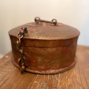 May include: A round, copper box with a hinged lid and a decorative chain and clasp. The box has an ornate design and a worn, aged patina.