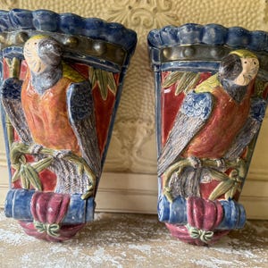 May include: Pair of ceramic wall sconces, each featuring a parrot design. The sconces have a blue scalloped top, a red background, and a parrot with orange, yellow, and blue plumage. The base is a combination of blue and pink with a decorative bow.