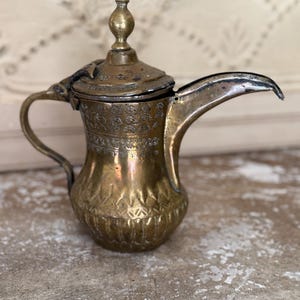 May include: Antique brass coffee pot with a curved spout and ornate detailing. The pot features a small handle and a decorative lid with a finial. The brass has a warm, aged patina, suggesting its vintage origin. A classic piece for display.