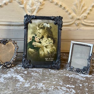May include: Three decorative metal picture frames. One heart-shaped frame with rose detailing, one rectangular frame with ornate scrollwork containing a vintage-style portrait of a girl holding flowers, and one rectangular frame with rose accents.