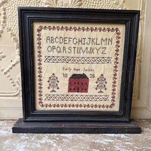 May include: Framed cross-stitch sampler with a black wooden frame. The sampler features the alphabet, a house, trees, and the text "Emily Hope Smithe 1826". The stitching is in black, red, and tan colors.