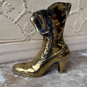May include: A gold-colored, vintage-style boot-shaped decorative item. The boot features a high heel, button details, and a folded-over top. The metallic finish gives it a shiny appearance, suggesting it's made of brass or a similar material.