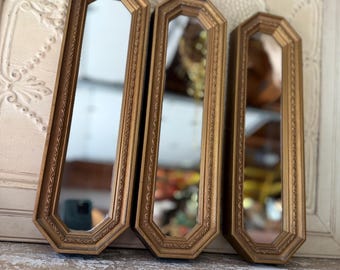 May include: Three vertical mirrors with elongated octagonal shapes and metallic gold frames. Each frame features a textured, beaded edge detail surrounding the reflective glass surface. The mirrors are arranged side by side in a row.