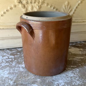 May include: A brown ceramic crock with a cylindrical shape and two small handles. The crock has a light brown interior and a darker brown exterior with a glossy finish. The crock is likely used for food storage.