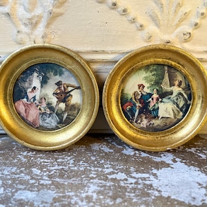 May include: Two circular wall plaques featuring ornate gold frames. The left plaque depicts a man playing a lute for two seated figures in a garden. The right plaque shows a group of three figures in period clothing gathered in an outdoor setting.