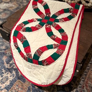 May include: A quilted table runner with a white background and a double wedding ring design in red and green. The runner has a red border and is placed on a wooden surface.