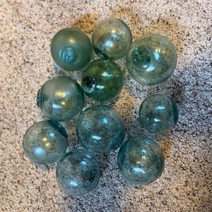 May include: A collection of ten glass fishing floats in varying shades of green and blue. The spheres have a textured, aged appearance, with visible imperfections and bubbles within the glass. They are arranged on a neutral-toned surface.