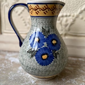 May include: A ceramic pitcher with a blue handle and spout. The pitcher is decorated with blue flowers and green leaves on a speckled gray background. The rim features a yellow and brown geometric pattern.