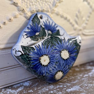 May include: A small, ceramic wall pocket with a blue and white floral design. The pocket features three large blue flowers with white centers and green leaves. The pocket is a light blue color and has a hole for hanging.