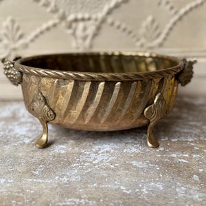 May include: A round brass bowl featuring a textured, diagonal ribbed pattern on the exterior, a twisted rope-style rim, and two decorative handles shaped like clusters of grapes. The bowl is supported by three ornate, curved metal feet.